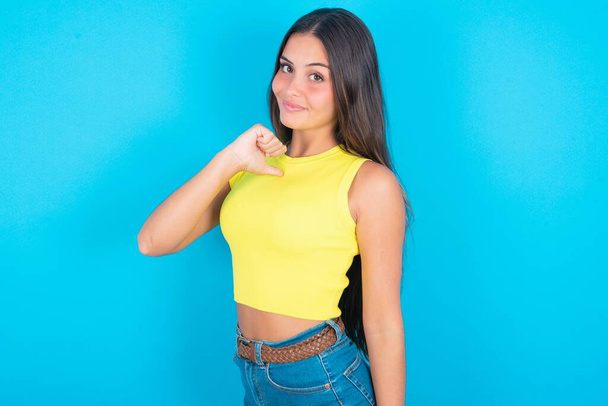 Closeup of cheerful beautiful brunette woman wearing yellow tank top over blue background looks joyful, satisfied and confident, points at himself with thumb. - Photo, Image