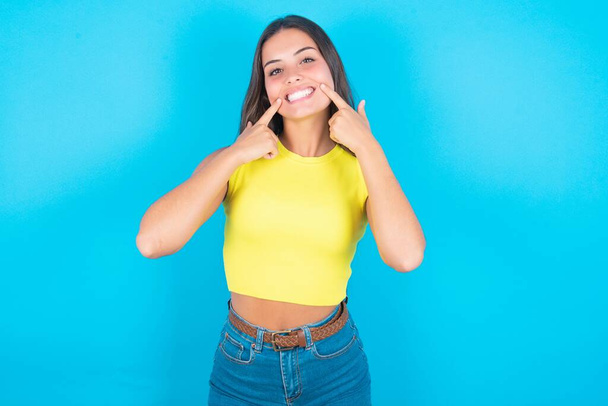 Erős, egészséges, egyenes, fehér fogak. Közelkép portré boldog szép barna nő visel sárga tartály tetején kék háttér sugárzó mosollyal mutató tökéletes tiszta fehér fogak. - Fotó, kép