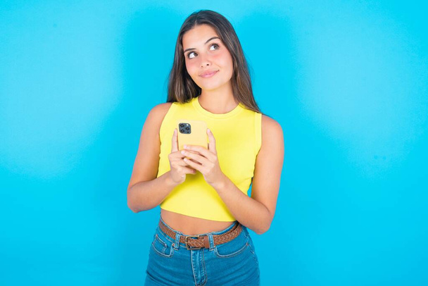 Portret pięknej brunetki noszącej żółtą bluzkę na niebieskim tle z marzycielskim wyglądem, myślącej trzymając smartfona. Próbuje napisać wiadomość.. - Zdjęcie, obraz