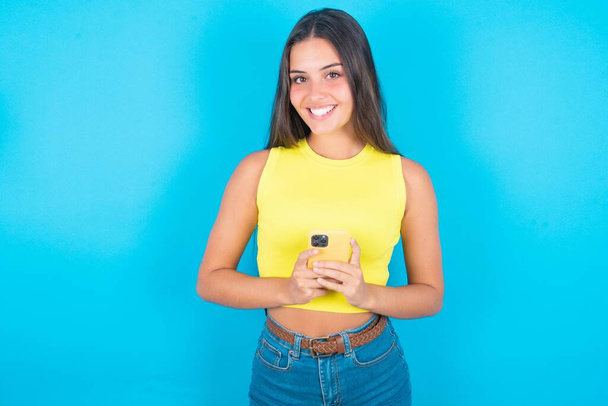 Lächelnde schöne brünette Frau mit gelbem Tanktop vor blauem Hintergrund freundlich und glücklich mit Handy Selfie im Spiegel. - Foto, Bild
