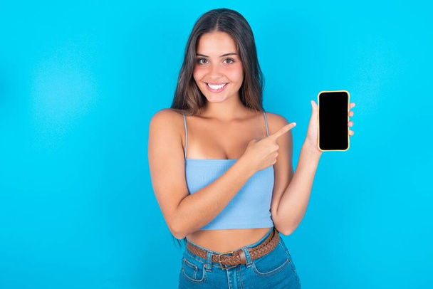 Uśmiechnięta biała brunetka ubrana w niebieską bluzkę ze zbiornikiem pokazującą i wskazującą na pusty ekran telefonu na niebieskim tle - Zdjęcie, obraz
