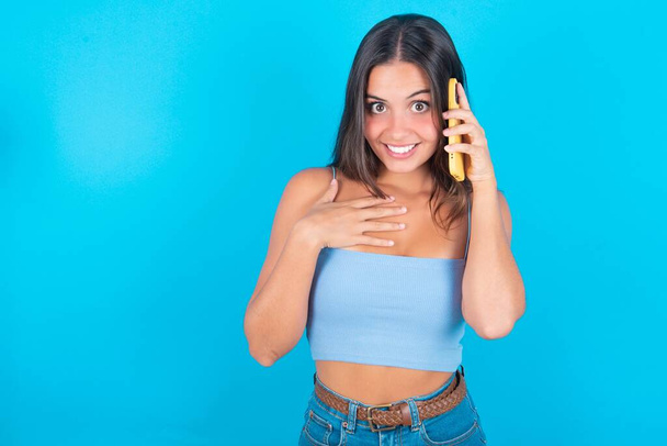 glimlachende jonge brunette vrouw dragen blauwe tank top over blauwe achtergrond gesprekken via mobiele telefoon, geniet van aangename groot gesprek. - Foto, afbeelding