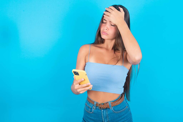 Verstoorde Kaukasische brunette vrouw draagt blauwe tank top maakt gezicht palm als vergeten over iets belangrijks houdt mobiele telefoon - Foto, afbeelding