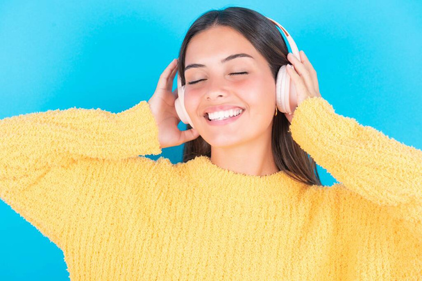 brunette woman with closed eyes wearing yellow sweater smiles broadly, listens favorite music track via wireless headphones  - Foto, Imagen
