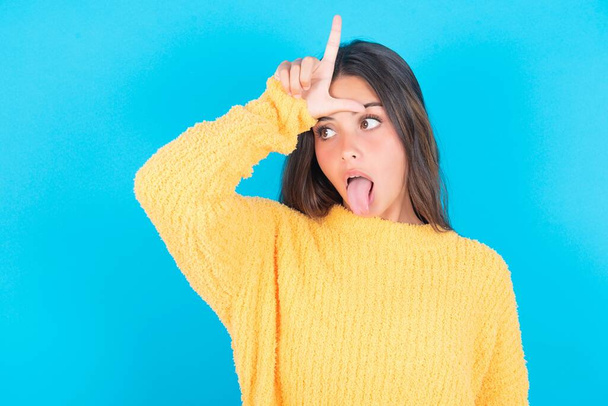 Funny brunette woman wearing yellow sweater makes loser gesture mocking at someone sticks out tongue - Photo, Image