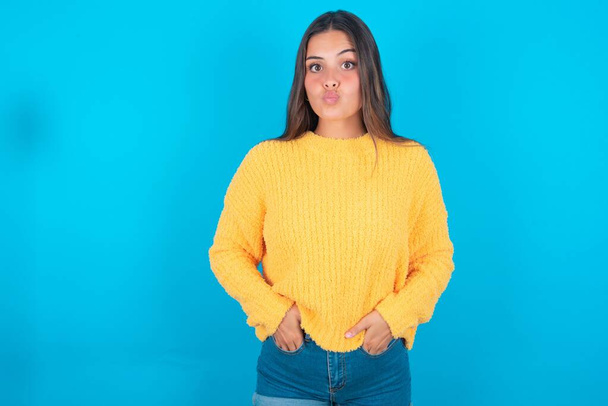 Портрет милой юной брюнетки в жёлтом свитере на синем фоне, посылающий воздушный поцелуй - Фото, изображение