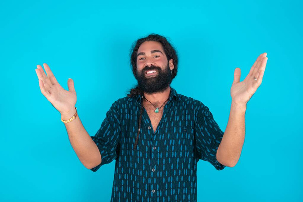 Веселий кавказький чоловік з бородою в синій сорочці на синьому фоні робить вітальний жест, піднімаючи руки над головою. - Фото, зображення