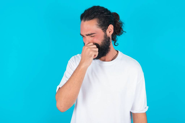 Homem caucasiano com barba vestindo camiseta branca sobre fundo azul segurando o nariz por causa de um mau cheiro. - Foto, Imagem