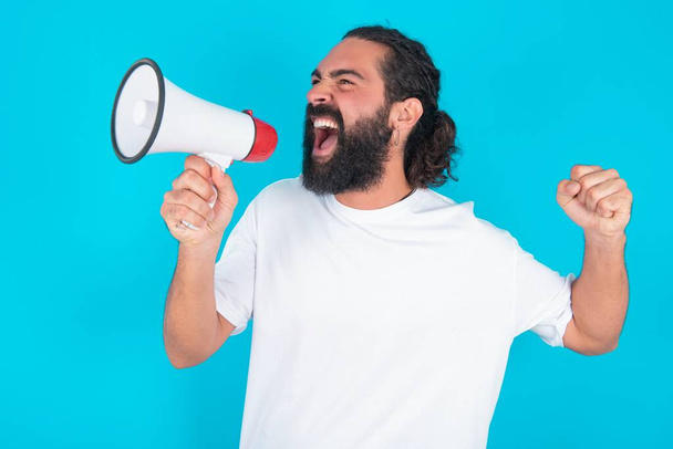Běloch s vousy v bílém tričku přes modré pozadí komunikuje hlasitě křičí drží megafon, vyjadřuje úspěch a pozitivní koncept, nápad pro marketing nebo prodej. - Fotografie, Obrázek
