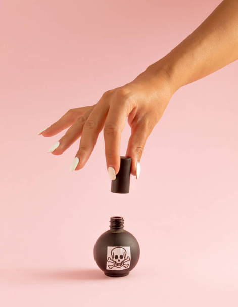 Mano de mujer con uñas rojas y veneno en botella negra sobre fondo rosa pastel. Halloween idea espeluznante surrealista. - Foto, imagen