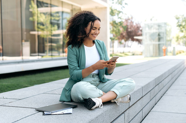 Happy young mixed race successful curly haired business woman, in elegant wear, holding smart phone outdoors, using gadget for online messaging with friends, checking email, browsing websites, smiles - Foto, Imagem