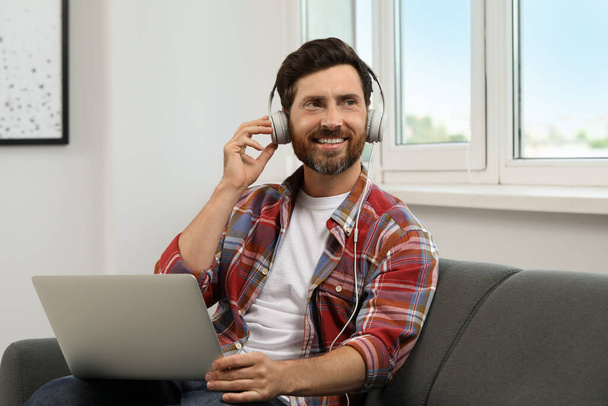 Smiling bearded man with headphones and laptop on sofa indoors - Valokuva, kuva