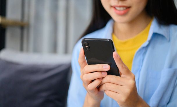 Hymyilevä nuori aasialainen nainen matkapuhelimella tarkistaa sosiaalisen median, sovellus pelaa peliä, ostoksia verkossa, tilaaminen toimitus rentoutua istuessaan sohvalla kotona kannettavan tietokoneen  - Valokuva, kuva