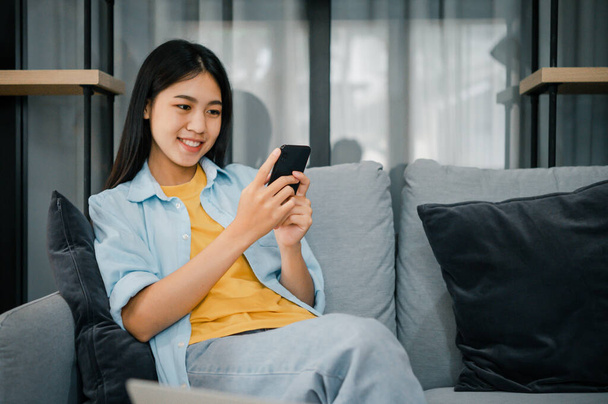 Усміхнена молода азіатка, використовуючи мобільний телефон, перевіряє соціальні медіа, додаток, грає в гру, купує онлайн, замовляє доставку відпочити, сидячи на дивані вдома з ноутбуком
  - Фото, зображення