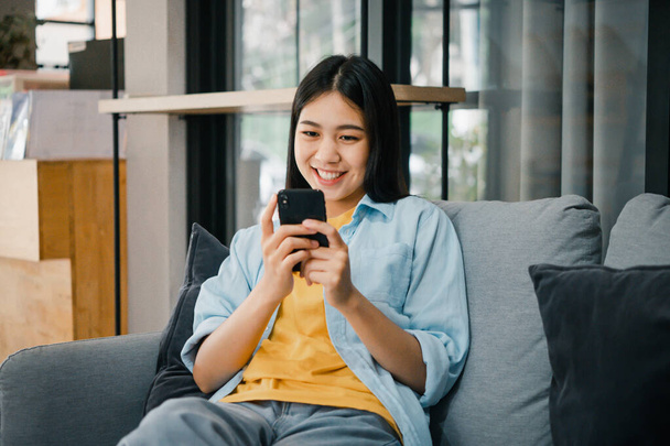 Sonriente joven mujer asiática utilizando el teléfono móvil comprobación de las redes sociales, aplicación de juego, compras en línea, la entrega de pedidos relajarse sentado en un sofá en casa con ordenador portátil  - Foto, Imagen