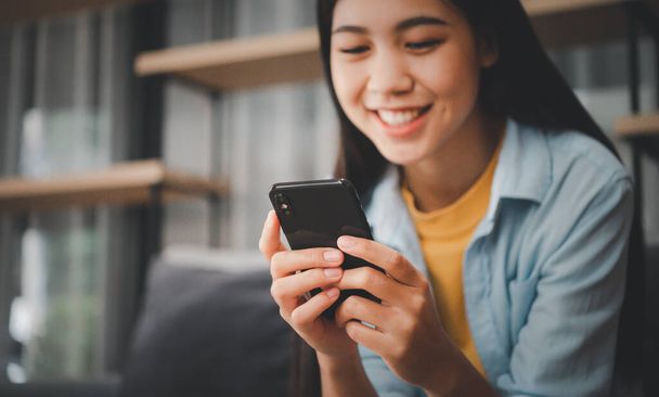 携帯電話のチェックを使用して若いアジアの女性を笑顔ソーシャルメディア、アプリゲームをプレイ、オンラインショッピング、ラップトップコンピュータで自宅でソファに座っている間に配達をリラックス注文  - 写真・画像