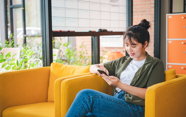 Smiling young asian woman using mobile phone checking social media,app playing game, shopping online, ordering delivery relax while sitting on a couch at cafe with laptop computer  - Photo, Image