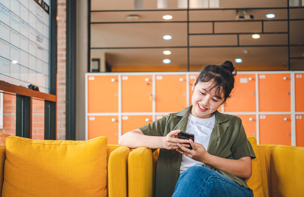 Hymyilevä nuori aasialainen nainen matkapuhelimella tarkistaa sosiaalisen median, sovellus pelaa peliä, ostoksia verkossa, tilaaminen toimitus rentoutua istuessaan sohvalla kahvilassa kannettavan tietokoneen  - Valokuva, kuva