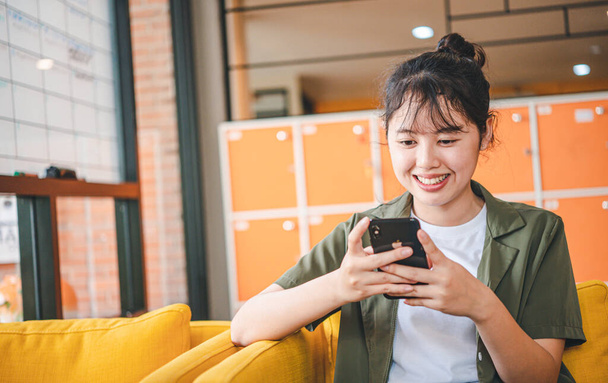 Lächelnde junge Asiatin, die mit dem Handy soziale Medien checkt, App spielt, online einkauft, Lieferung bestellt, entspannt auf der Couch im Café mit Laptop sitzt  - Foto, Bild