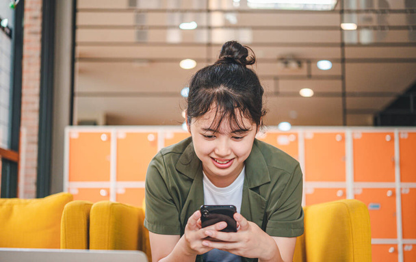 Lächelnde junge Asiatin, die mit dem Handy soziale Medien checkt, App spielt, online einkauft, Lieferung bestellt, entspannt auf der Couch im Café mit Laptop sitzt  - Foto, Bild