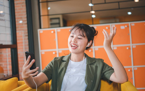 Lachende jonge Aziatische vrouw met behulp van mobiele telefoon controleren van sociale media, app spel spelen, online winkelen, bestellen levering ontspannen terwijl zitten op een bank in cafe met laptop computer  - Foto, afbeelding