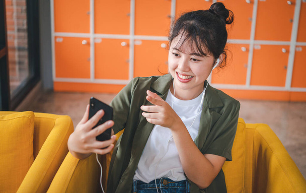 携帯電話のチェックを使用して若いアジアの女性を笑顔ソーシャルメディア、アプリゲームをプレイ、オンラインショッピング、ラップトップコンピュータでカフェでソファに座っている間に配達をリラックスさせる  - 写真・画像