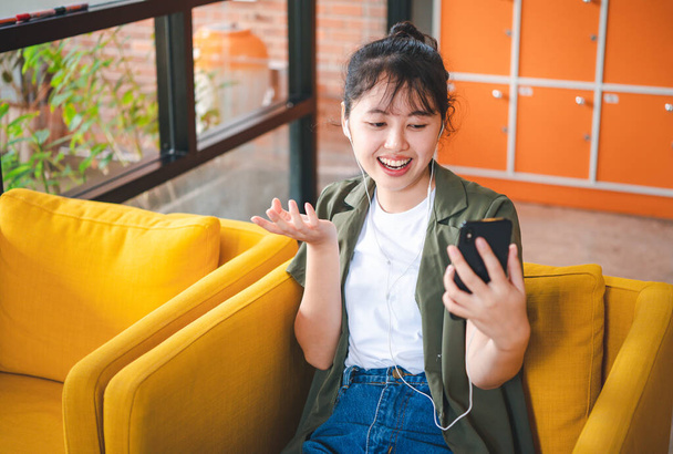 Улыбающаяся молодая азиатка, пользующаяся мобильным телефоном, проверяющая социальные сети, играющая в игры, покупающая онлайн, заказывающая доставку отдыхает, сидя на диване в кафе с ноутбуком  - Фото, изображение