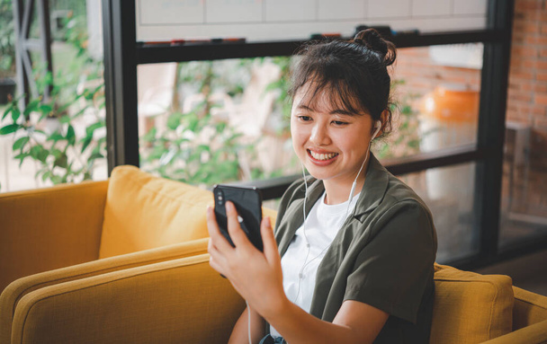 Sonriendo joven mujer asiática utilizando el teléfono móvil comprobación de las redes sociales, aplicación de juego, compras en línea, la entrega de pedidos relajarse sentado en un sofá en la cafetería con ordenador portátil  - Foto, Imagen