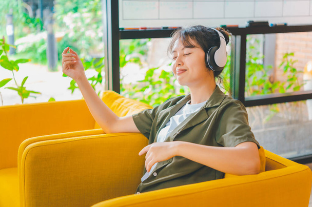 teenage woman wearing headphones sits on her phone while listening to music, playing games, ordering online on the sofa comfortably in a cafe. - Photo, Image