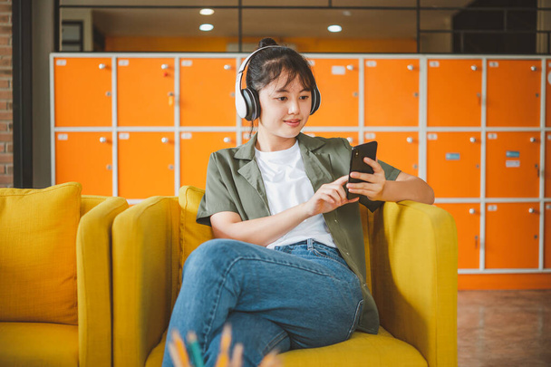 tiener vrouw draagt koptelefoon zit op haar telefoon tijdens het luisteren naar muziek, het spelen van games, online bestellen op de bank comfortabel in een cafe. - Foto, afbeelding