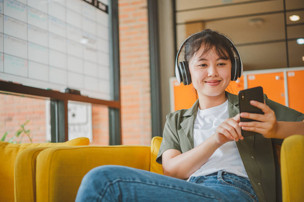teini-ikäinen nainen, jolla on kuulokkeet, istuu puhelimessaan kuunnellen musiikkia, pelaten pelejä, tilaamalla verkossa sohvalla mukavasti kahvilassa.. - Valokuva, kuva