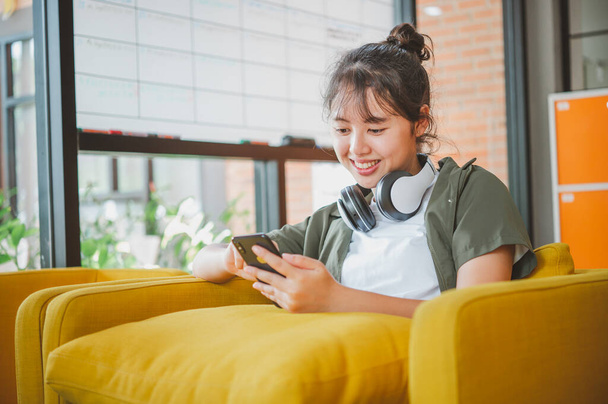 mujer adolescente con auriculares se sienta en su teléfono mientras escucha música, juega juegos, ordena en línea en el sofá cómodamente en un café. - Foto, Imagen