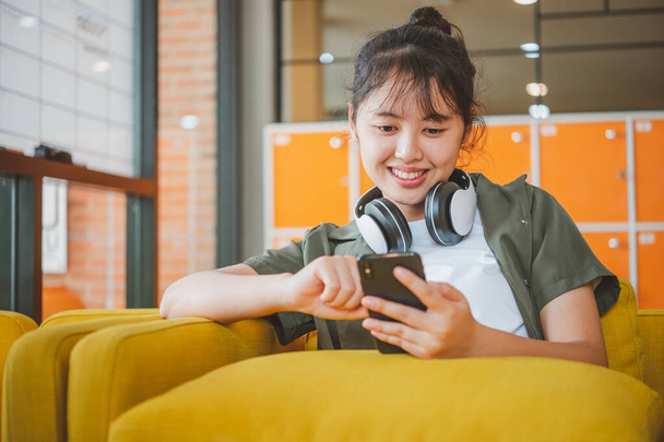 Teenager-Frau mit Kopfhörer sitzt auf ihrem Handy, hört Musik, spielt Spiele und bestellt bequem online auf dem Sofa in einem Café. - Foto, Bild