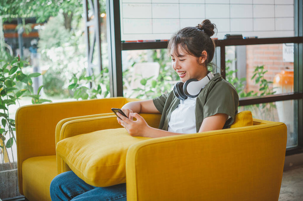 mulher adolescente usando fones de ouvido senta-se em seu telefone enquanto ouve música, jogar jogos, encomendar on-line no sofá confortavelmente em um café. - Foto, Imagem