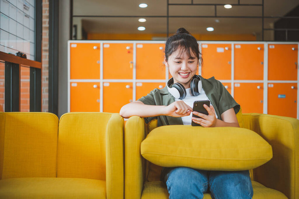 teini-ikäinen nainen, jolla on kuulokkeet, istuu puhelimessaan kuunnellen musiikkia, pelaten pelejä, tilaamalla verkossa sohvalla mukavasti kahvilassa.. - Valokuva, kuva