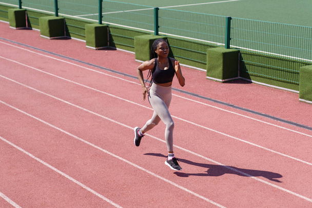Young black sportswoman enjoys running on red track around stadium to improve technique and increase endurance. African American woman with long braids prepares for upcoming marathon on hot summer day - Foto, Imagem