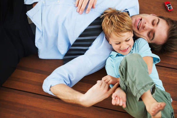 Σταμάτα μπαμπά, με γαργαλάς. Ένα χαριτωμένο αγοράκι να γελάει στο πάτωμα ενώ τον γαργαλάει ο νεαρός πατέρας του. - Φωτογραφία, εικόνα