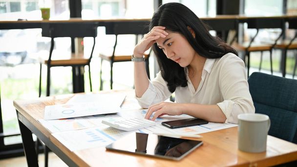 Junge asiatische Geschäftsfrau oder Buchhalterin, gestresst und nachdenklich, arbeitet fernab von einem Café und konzentriert sich auf ihren Bericht. - Foto, Bild