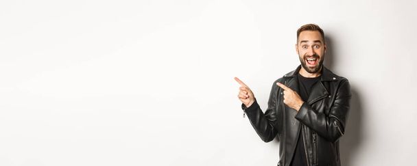 Άνδρας με αυτοπεποίθηση με μαύρο δερμάτινο μπουφάν, που δείχνει τα δάχτυλα αριστερά στην προσφορά promo, που δείχνει το λογότυπο, λευκό φόντο. - Φωτογραφία, εικόνα