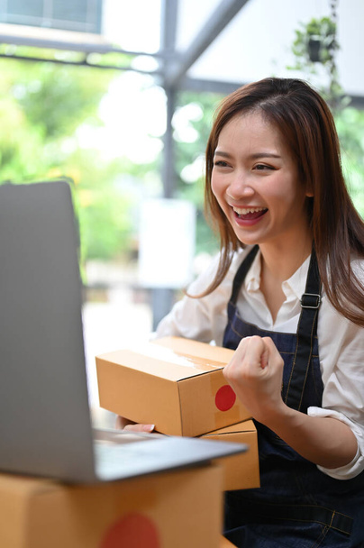 Portret, Happy jonge Aziatische vrouwelijke e-commerce ondernemer op zoek naar laptop scherm, enthousiast over haar online verkoop. Online bedrijfsconcept. - Foto, afbeelding