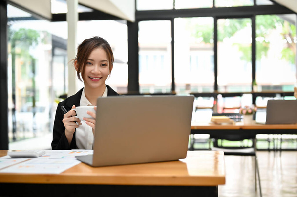 Atraktivní mladá asijská podnikatelka nebo žena kancelář zaměstnanec dálkově pracuje v kavárně, s kávou, zatímco pracuje své úkoly na svém přenosném notebooku. - Fotografie, Obrázek