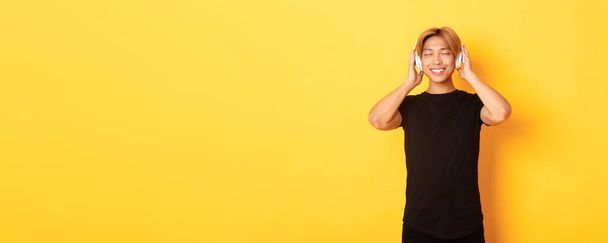 Relaxed and pleased handsome asian guy close eyes from satisfaction while listening music in wireless headphones, standing yellow background. - Photo, Image