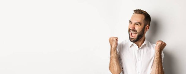 Close-up of successful caucasian man rejoicing, making fist pump and shouting of joy, looking left, winning, standing over white background. - 写真・画像