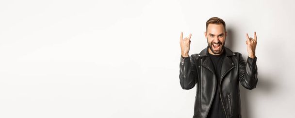 Homem adulto fresco em jaqueta de couro preto, mostrando rock em gesto e língua, apreciando festival de música, de pé sobre fundo branco. - Foto, Imagem