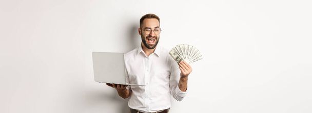 Business and e-commerce. Confident businessman showing how work online, winking, holding money and laptop, standing over white background. - Photo, Image