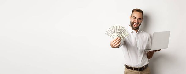 Business and e-commerce. Happy successful businessman bragging with money, working on laptop online, standing over white background. - Photo, Image