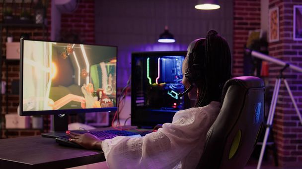 African american gamer girl using pc gaming setup having a good time playing multiplayer online action game in home living room. Woman streaming first person shooter while talking in headset. - 写真・画像