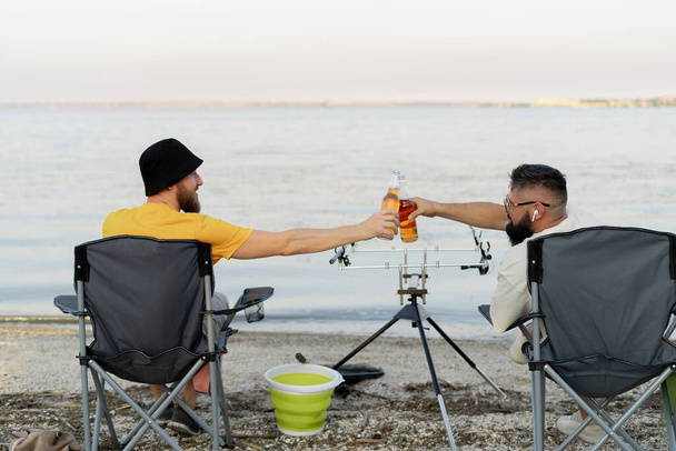 Twee vissers zitten samen met bier tijdens het vissen op het meer in de zomerochtend. Vrijetijdsbesteding en mensen - Foto, afbeelding
