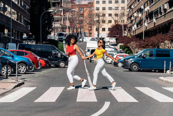 Pretty african american young women holding hands and walking in zebra crossing in the city and having fun. - Foto, Bild