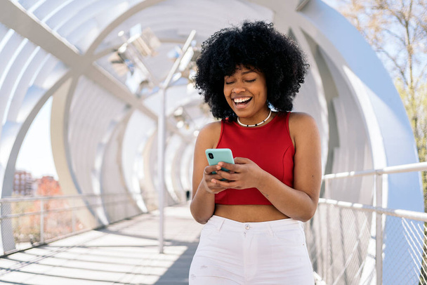 Cool fiatal fekete nő afro haj mosolyog, és használja a mobiltelefon az utcán. - Fotó, kép